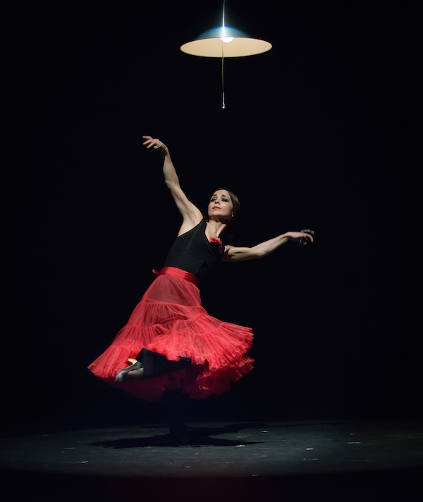 Perciet in a in a red full flamenco skirt extends her arms over her head while one foot in the air